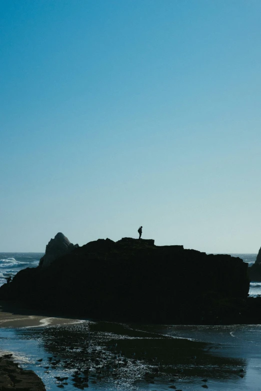 a person standing on top of a rock near the ocean, silhouetted, rock formations, a long-shot, zoomed out