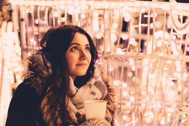 a woman holding a cup of coffee in front of christmas lights, by Julia Pishtar, pexels contest winner, art nouveau, soft pink lights, in an igloo, clara oswald, portrait of a beautiful