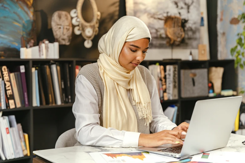 a woman sitting at a desk using a laptop computer, inspired by Maryam Hashemi, trending on pexels, hurufiyya, white hijab, studious, decorative, ethnic origin