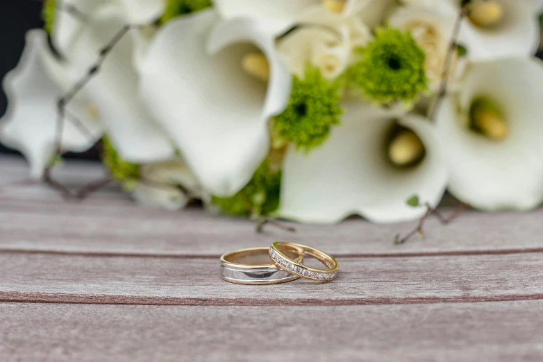 two wedding rings sitting next to a bouquet of flowers, by Sylvia Wishart, unsplash, on wooden table, gold and white, platinum jewellery, sit on a bench