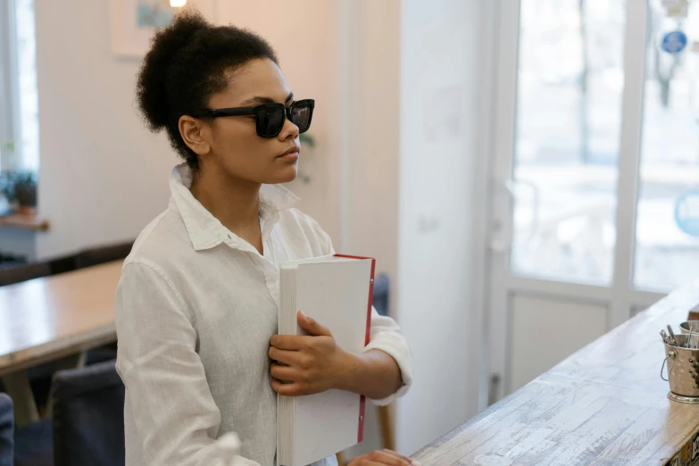 a woman standing in front of a counter holding a book, trending on unsplash, fantastic realism, implanted sunglasses, tessa thompson inspired, standing in class, queer woman