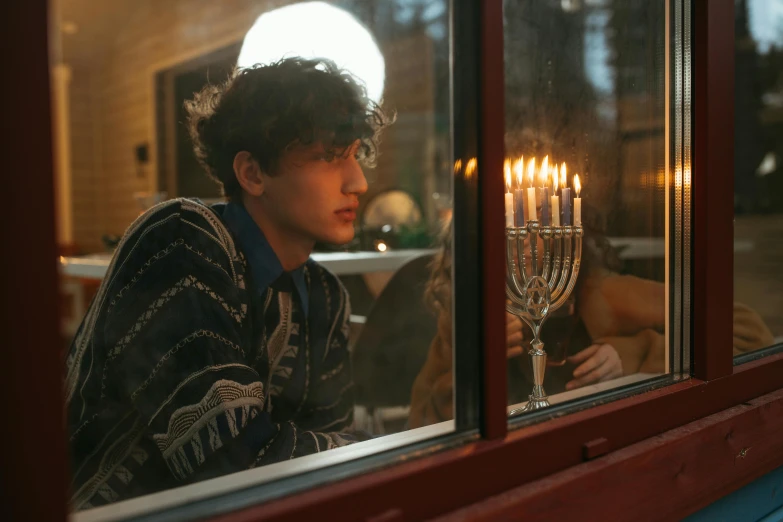 a boy looking out a window at a lit menorah, an album cover, pexels, male teenager, movie still 8 k, ignant, curls on top of his head