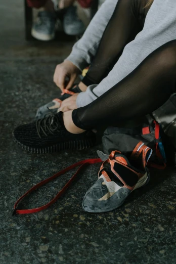 a woman sitting on the ground tying up her shoes, by Louisa Matthíasdóttir, trending on unsplash, dark grey and orange colours, wearing adidas clothing, non-binary, rugged details