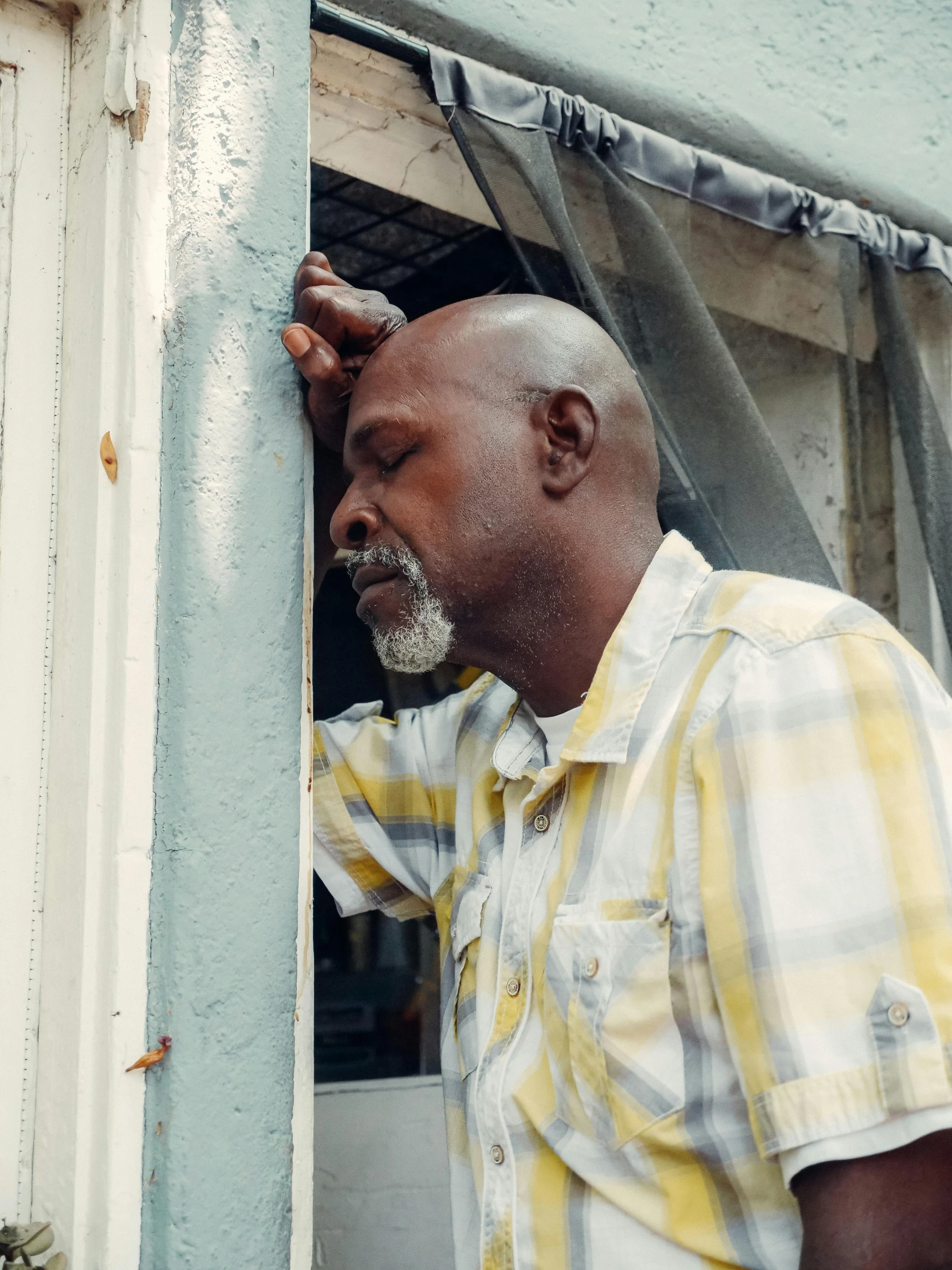 a man in a yellow and white shirt looking out of a window, a photo, grieving, in louisiana, leaning on door, 2019 trending photo