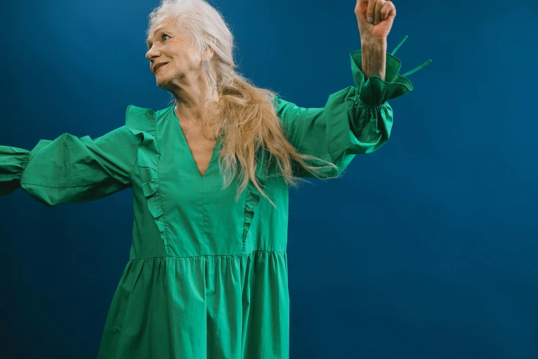 a woman in a green dress standing in front of a blue background, pexels contest winner, renaissance, elderly, playful pose of a dancer, with long white hair, still from a music video