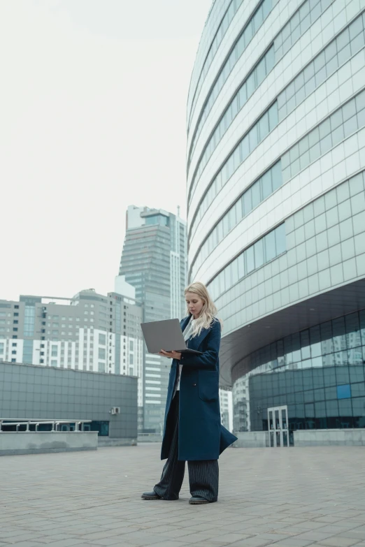 a woman standing in front of a building with a laptop, tall buildings, russian academic, technology cloak, trending