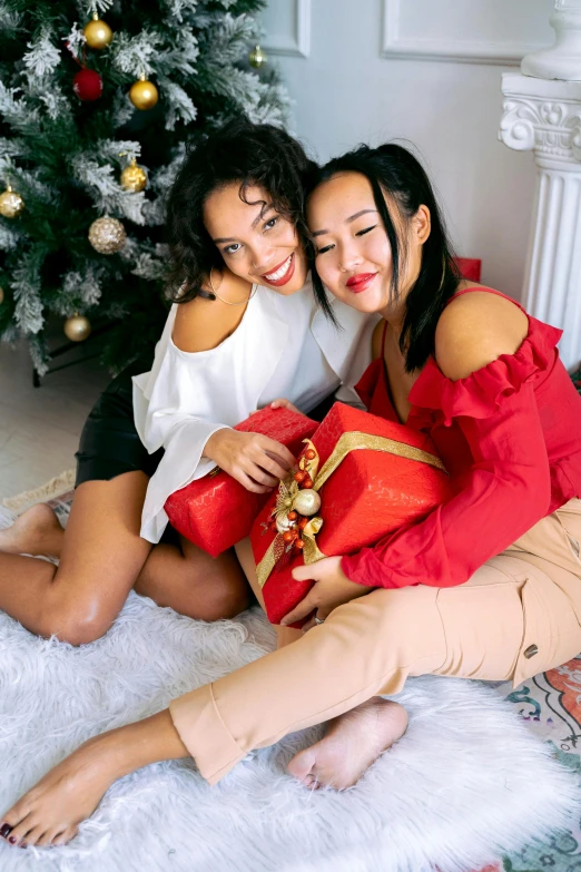 a couple of women sitting next to a christmas tree, influencer, woman holding another woman, wearing red, holding gift