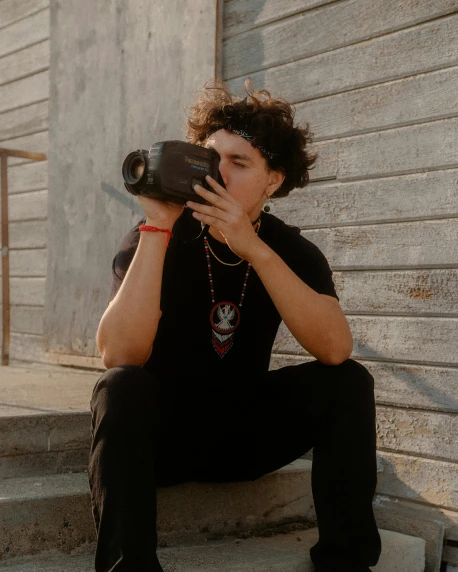 a young man sitting on the steps of a building, by Cosmo Alexander, pexels contest winner, video art, holding a camera, curly haired, he wears dark visors, ariel perez