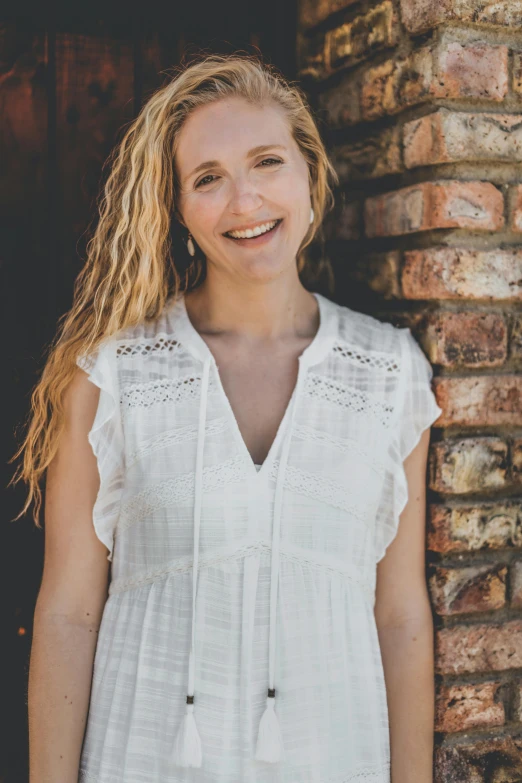 a woman standing in front of a brick wall, by Carey Morris, wearing a white folkdrakt dress, smiling down from above, curly blonde hair | d & d, cameron diaz portrait