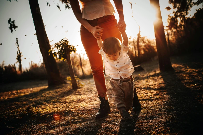 a woman holding the hand of a small child, by Jesper Knudsen, pexels, walking at the park, sun behind him, thumbnail, brown