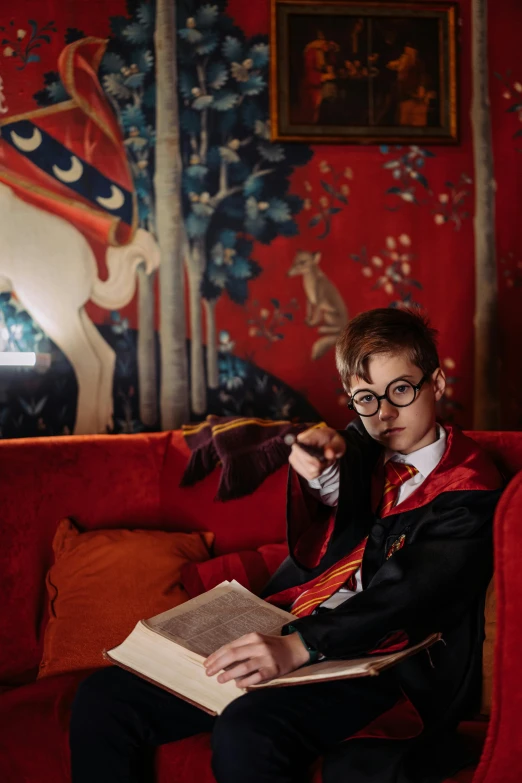 a man sitting on a red couch reading a book, inspired by Cecil Beaton, maximalism, magic school uniform, still from harry potter, young boy, wearing headmistress uniform