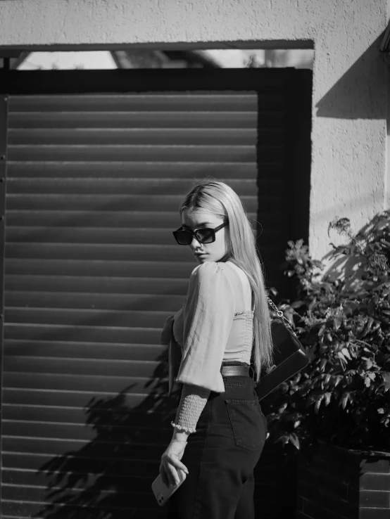 a woman standing in front of a garage door, a black and white photo, by Zofia Stryjenska, unsplash, wearing shades, young blonde woman, full body:: sunny weather::, anna nikonova aka newmilky