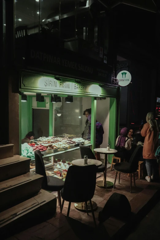 a group of people sitting at tables in front of a store, a picture, trending on unsplash, hurufiyya, very dark with green lights, cute bakery shop, background image