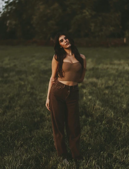 a woman standing in a field with trees in the background, an album cover, inspired by Elsa Bleda, trending on pexels, renaissance, brown pants, croptop, madison beer girl portrait, standing in a dimly lit room