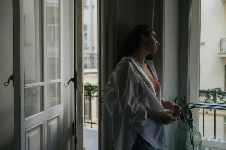 a woman standing on a balcony looking out a window, inspired by Elsa Bleda, pexels contest winner, clothed in white shirt, heartbroken, in her room, queer woman