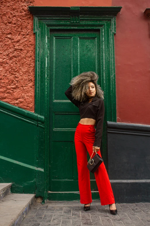 a woman standing in front of a green door, an album cover, by Julia Pishtar, trending on pexels, red gold and black outfit, red sweater and gray pants, harlem, pose model