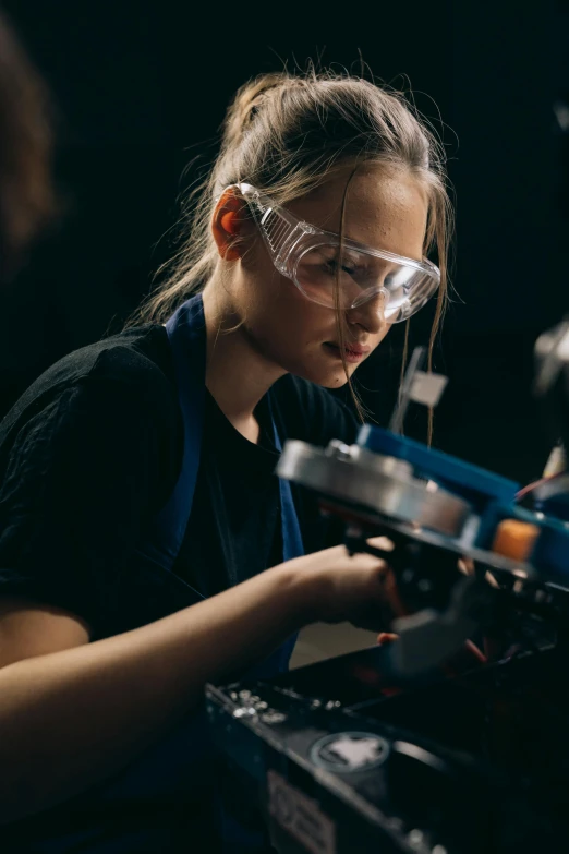 a woman working on a machine in a dark room, wearing goggles, aged 13, engineering bay, avatar image