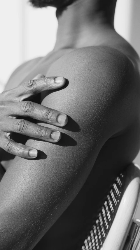 a man sitting in a chair with his hands on his chest, inspired by Max Dupain, unsplash, black young woman, close-up on legs, sunbathed skin, showing her shoulder from back