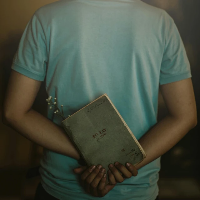 a person holding a book in their hands, an album cover, by Elsa Bleda, pexels, wearing a light blue shirt, his arms are behind his back, bible, grunge
