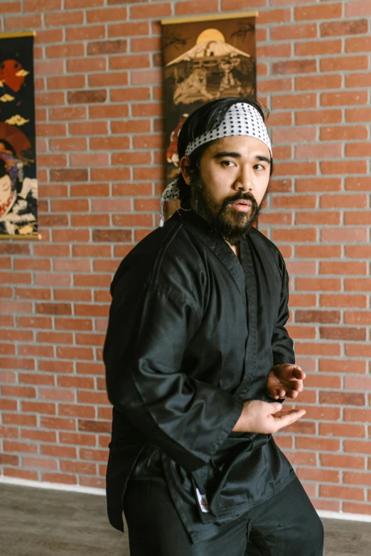 a man standing in front of a brick wall, a portrait, inspired by Kanō Sansetsu, reddit, in a dojo, performance, with a beard and a black shirt, bao pham