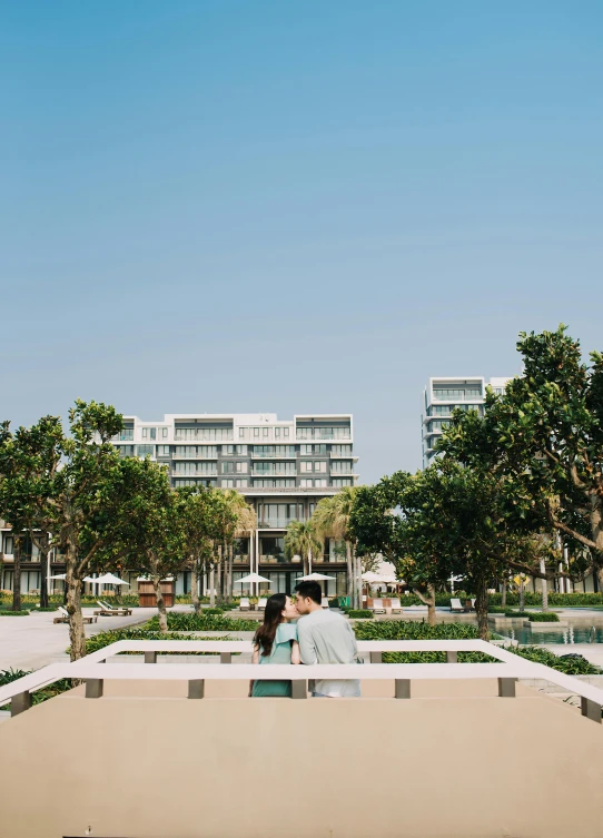 a couple sitting on a bench in front of a building, in a beachfront environment, in the center of the image, lush surroundings, raising between the buildings