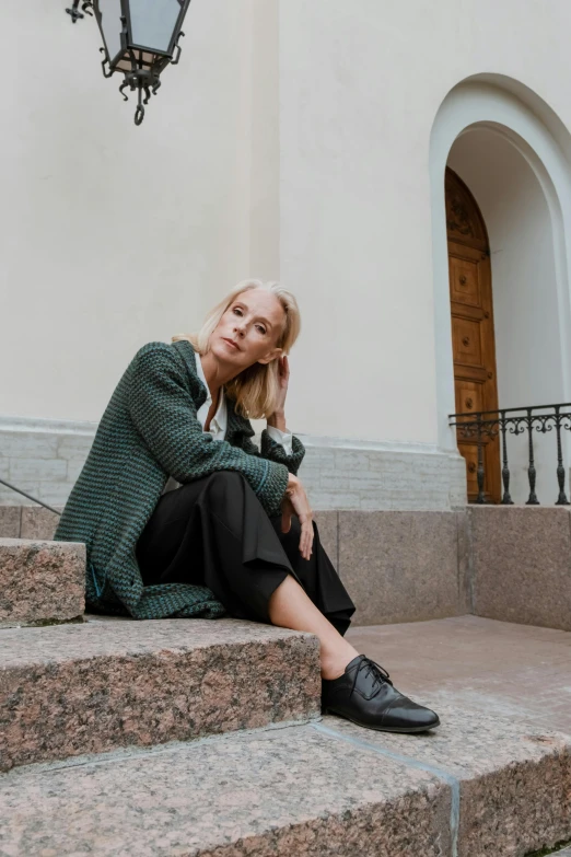 a woman sitting on the steps of a building, by Grytė Pintukaitė, trending on unsplash, wearing a cardigan, green and black colors, blonde swedish woman, thoughtful pose