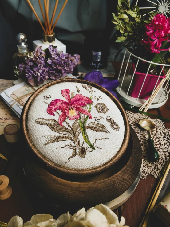 a table topped with a wooden bowl filled with flowers, a cross stitch, inspired by Maria Sibylla Merian, trending on pexels, orchid, circle iris detailed structure, album art, brown and magenta color scheme