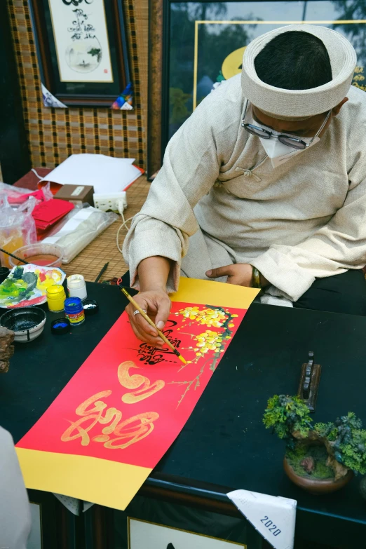 a man sitting at a table working on a painting, a silk screen, inspired by Wu Changshuo, islamic, ao dai, banner, red and yellow scheme