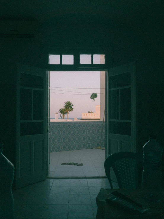 an open door leading to a patio with a table and chairs, by Youssef Howayek, lo-fi, low quality photo, palm trees in the background, dehazed image