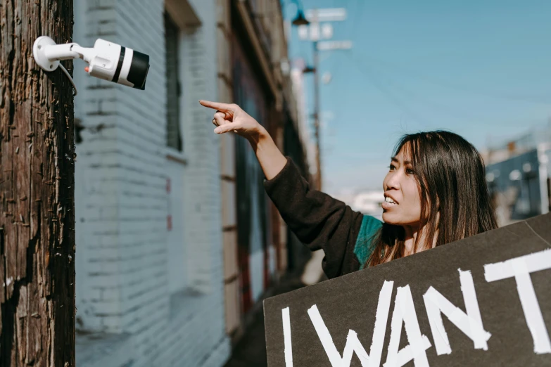 a woman holding a sign that says i want it, by Emma Andijewska, pexels contest winner, graffiti, camera angle looking up at her, protest, an asian woman, holding flask in hand