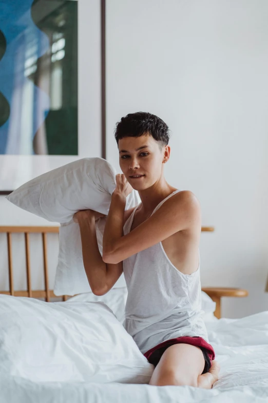 a man sitting on a bed holding a pillow, by Jessie Alexandra Dick, trending on unsplash, androgynous face, profile image, non binary model, sleek white