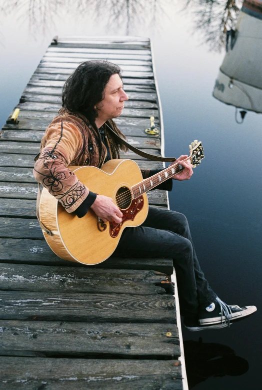 a man sitting on a dock playing a guitar, with his long black hair, linsey levendall, on a pale background, skin : tjalf sparnaay