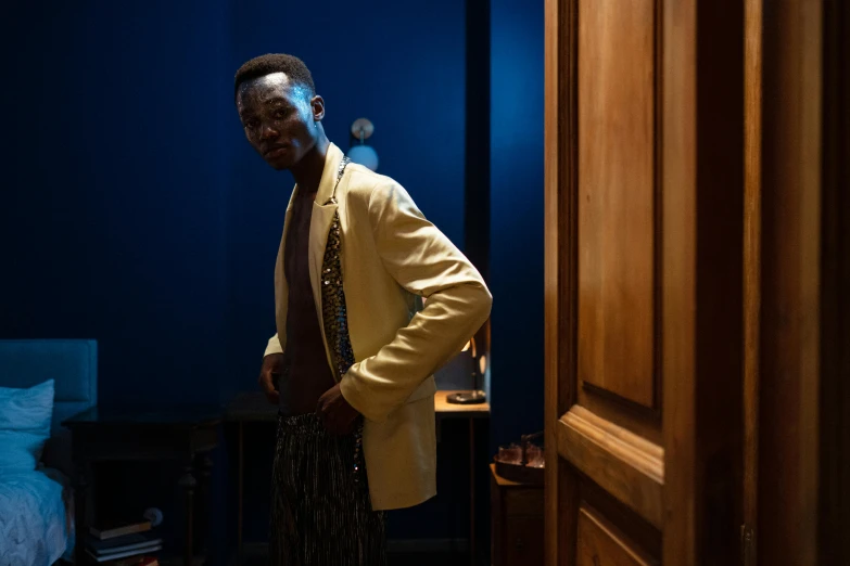 a man standing in a room next to a bed, an album cover, inspired by Barthélemy Menn, pexels contest winner, hyperrealism, blue gold suit, leaning on door, adut akech, movie still 8 k