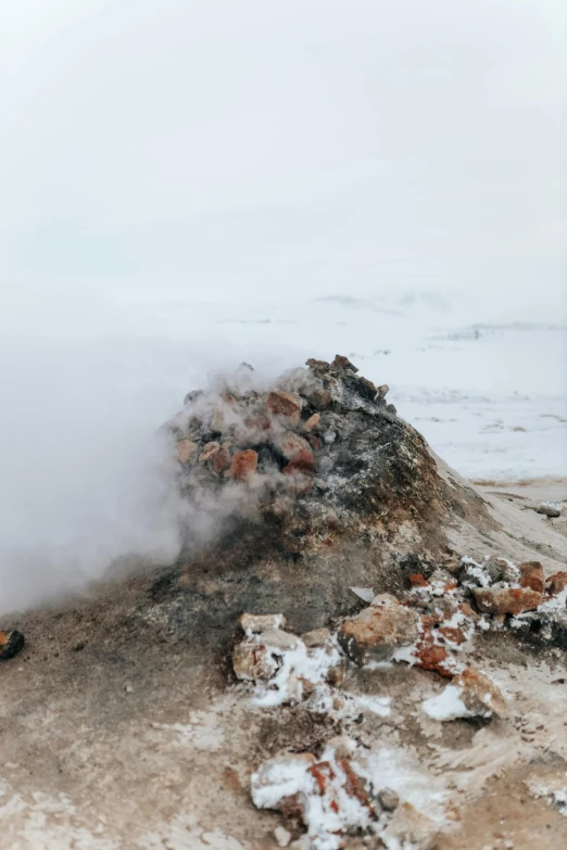 a man standing on top of a beach next to a pile of rocks, by Anna Haifisch, trending on unsplash, steaming food on the stove, sparse frozen landscape, reykjavik, exploding powder