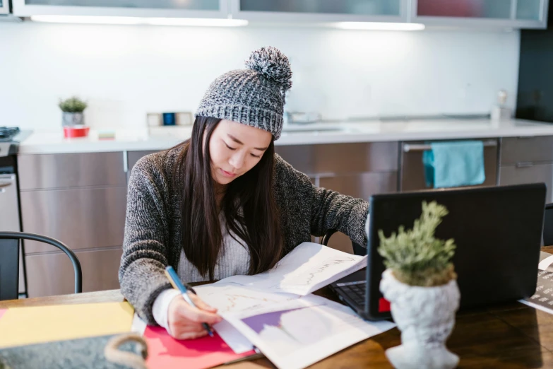 a woman sitting at a table working on a laptop, by Nicolette Macnamara, pexels contest winner, academic art, wearing a beanie, 9 9 designs, japanese, small and cosy student bedroom