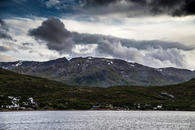 a body of water with mountains in the background, by Roar Kjernstad, pexels contest winner, hurufiyya, dark clouds above, settlement, panoramic, fan favorite