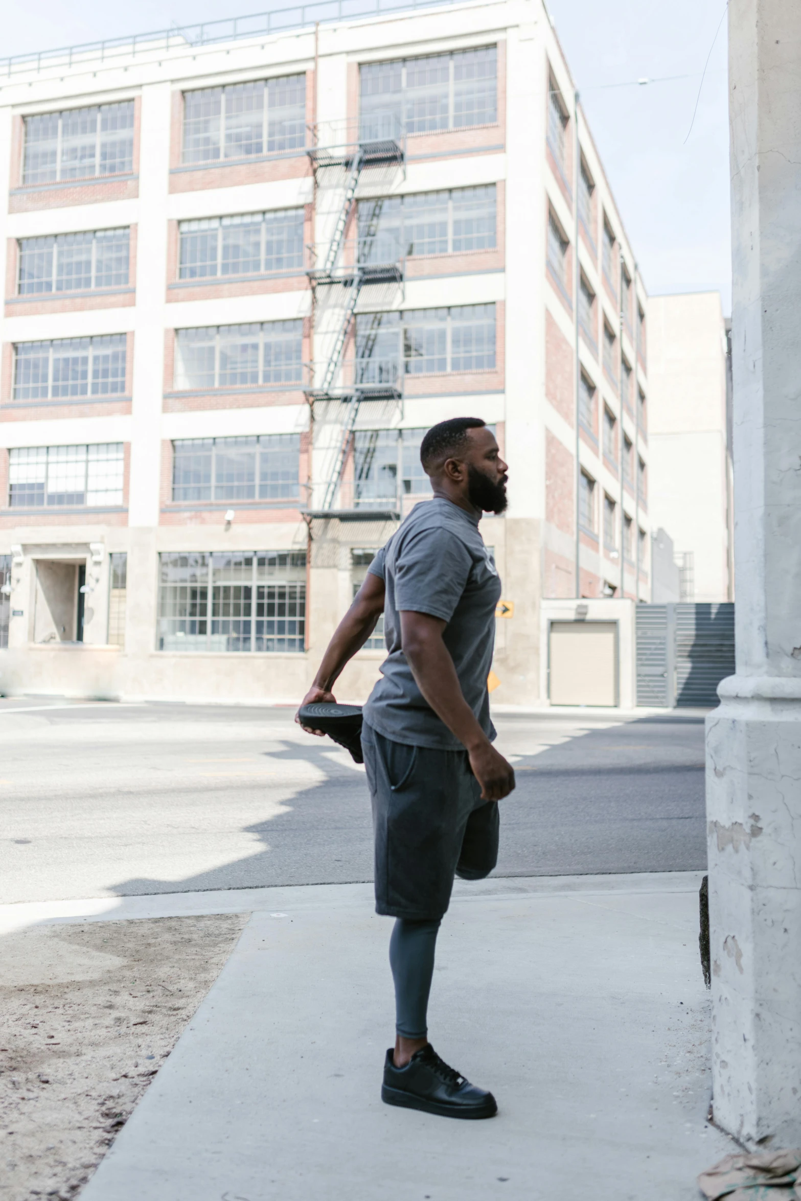 a man standing on a sidewalk in front of a building, working out, in gunmetal grey, black man, ponytail and beard