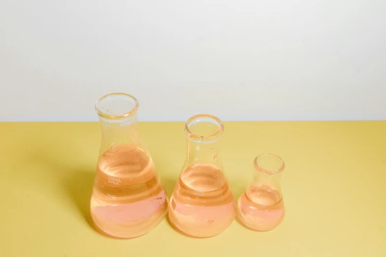 three glass vases filled with pink liquid on a yellow surface, unsplash, plasticien, wearing lab coat and a blouse, beakers, orange hue, small