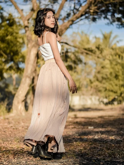 a woman walking down a dirt road next to a tree, by Lucia Peka, trending on pexels, renaissance, pleated skirt, posed in profile, stained dirty clothing, white and light-pink outfit