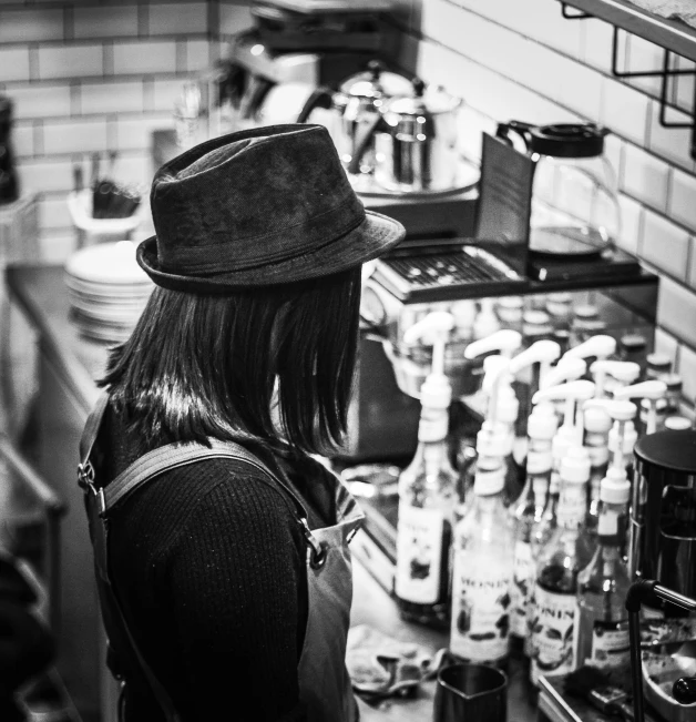 a black and white photo of a woman in a kitchen, by Adam Marczyński, pexels, buying beers in the british bar, character with a hat, coffee machine, monochrome:-2