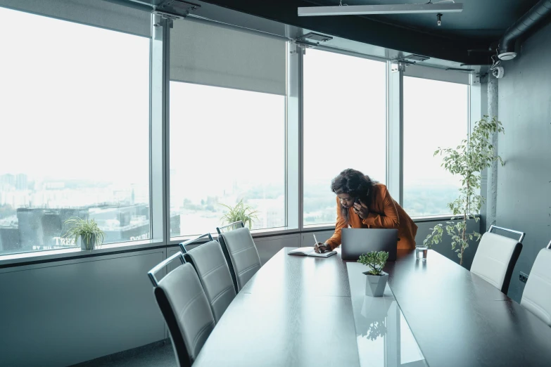 a woman sitting at a table working on a laptop, by Andries Stock, pexels contest winner, floor - to - ceiling windows, sad man, in a meeting room, worksafe. cinematic