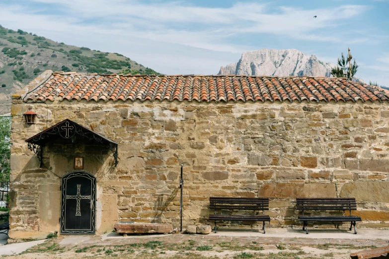 a stone building with two benches in front of it, by Pedro Álvarez Castelló, pexels contest winner, romanesque, behind a tiny village, background image, jesus alonso iglesias, freddy mamani silvestre facade