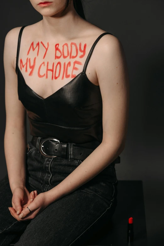 a woman with the words my body is a choice painted on her chest, an album cover, inspired by Nan Goldin, trending on pexels, feminist art, leather clothing, college, round-cropped, betty cooper