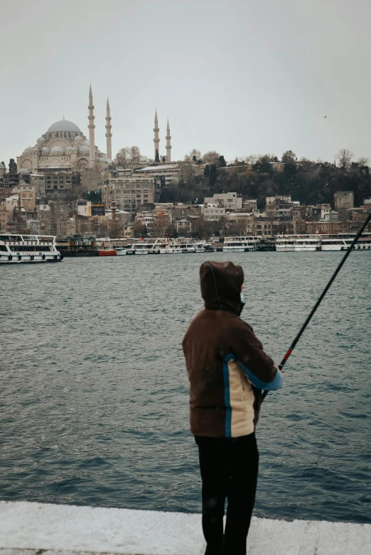 a man standing next to a body of water holding a fishing pole, a colorized photo, by irakli nadar, pexels contest winner, hurufiyya, istanbul, black domes and spires, a blond, gray skies