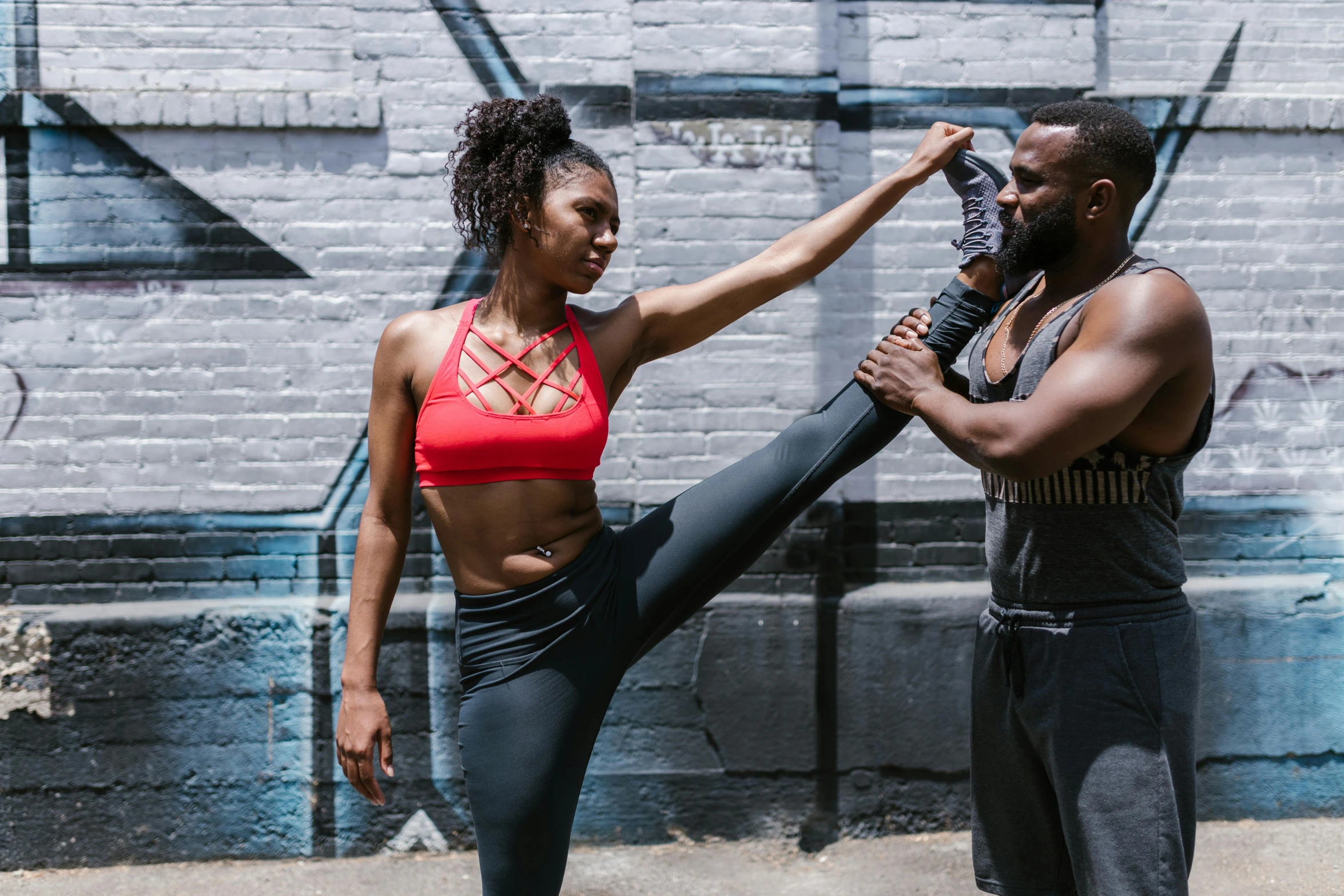 a man and a woman standing in front of a graffiti wall, by Nina Hamnett, pexels contest winner, black arts movement, doing splits and stretching, cropped red yoga short, athletic crossfit build, cast