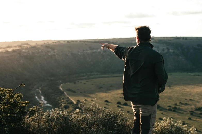 a man standing on top of a lush green hillside, by Lee Loughridge, pexels contest winner, happening, pointing, looking off into the sunset, lachlan bailey, subtle detailing