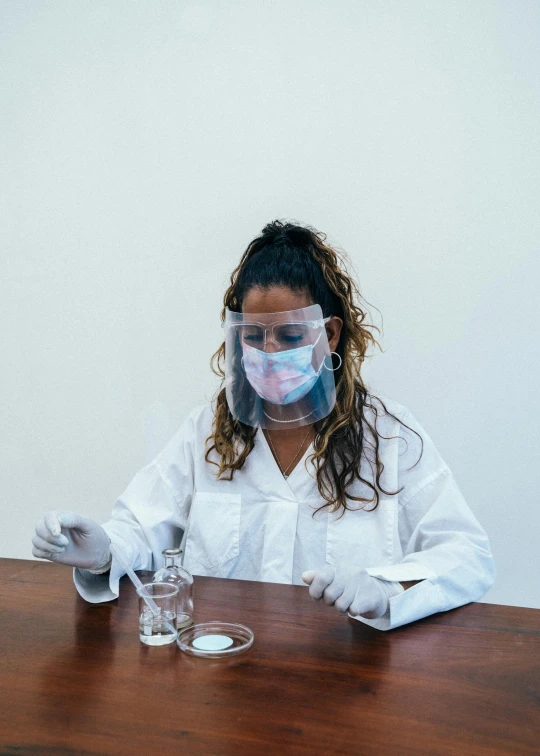 a woman in a lab coat sitting at a table, by Olivia Peguero, pexels, process art, wearing transparent glass mask, sao paulo, beakers, peru