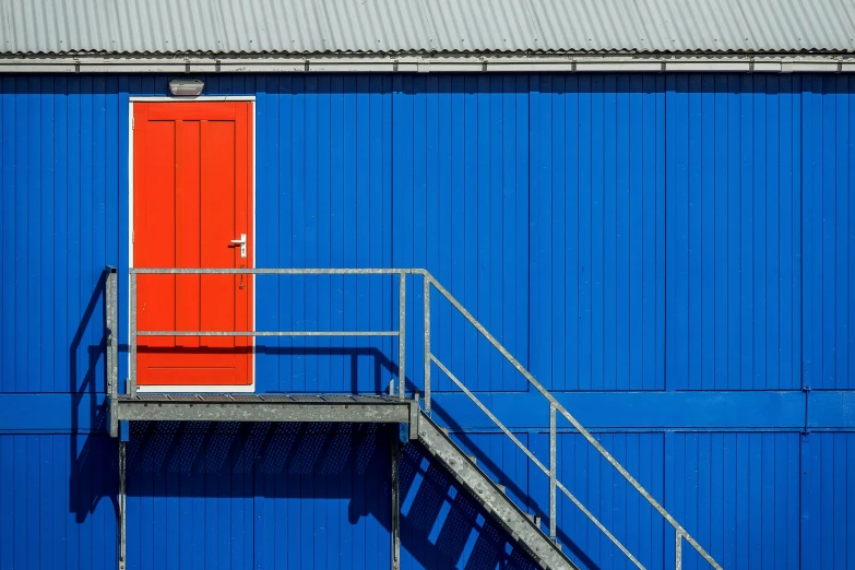 a blue building with a red door and stairs, by Sven Erixson, pexels contest winner, industrial colours, shed, some orange and blue, rolf klep