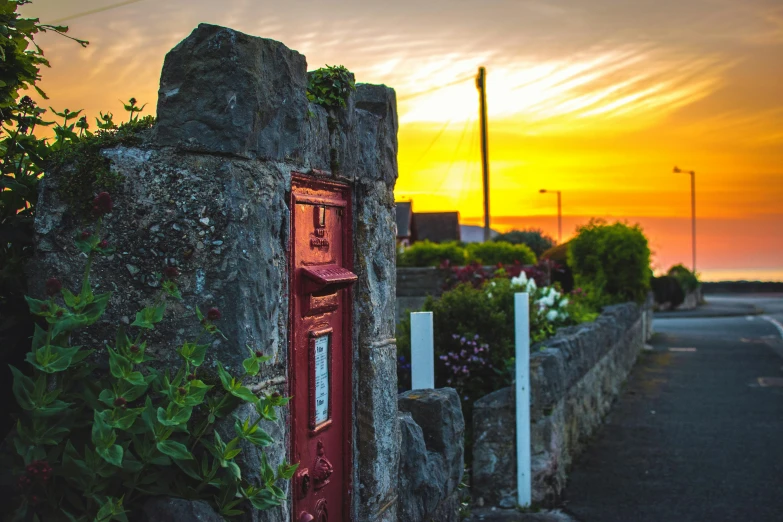 a red post box sitting on the side of a road, a picture, by Matt Stewart, pexels contest winner, private press, medieval coastal village, vibrant sunset, street of teal stone, an intricate