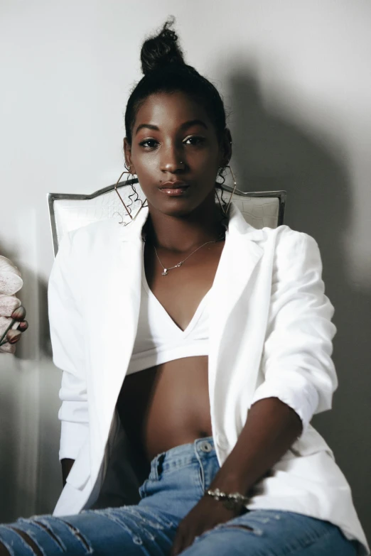 a woman in a white jacket sitting on a chair, inspired by Lily Delissa Joseph, trending on pexels, wearing a crop top, ( ( dark skin ) ), androgyny, bralette